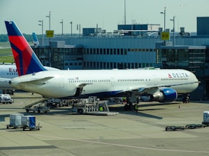 A large commercial airplane is stationed at an airport gate, connected via a jet bridge. The aircraft is a Delta Airlines plane, with visible airline branding. Surrounding the plane are various ground support vehicles, equipment, and personnel engaged in pre-flight or post-flight operations. The terminal building, with signage indicating gates, forms the backdrop.