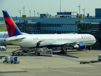 A large commercial airplane is stationed at an airport gate, connected via a jet bridge. The aircraft is a Delta Airlines plane, with visible airline branding. Surrounding the plane are various ground support vehicles, equipment, and personnel engaged in pre-flight or post-flight operations. The terminal building, with signage indicating gates, forms the backdrop.
