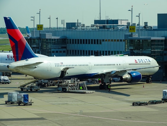 A large commercial airplane is stationed at an airport gate, connected via a jet bridge. The aircraft is a Delta Airlines plane, with visible airline branding. Surrounding the plane are various ground support vehicles, equipment, and personnel engaged in pre-flight or post-flight operations. The terminal building, with signage indicating gates, forms the backdrop.