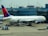 A large commercial airplane is stationed at an airport gate, connected via a jet bridge. The aircraft is a Delta Airlines plane, with visible airline branding. Surrounding the plane are various ground support vehicles, equipment, and personnel engaged in pre-flight or post-flight operations. The terminal building, with signage indicating gates, forms the backdrop.