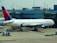 A large commercial airplane is stationed at an airport gate, connected via a jet bridge. The aircraft is a Delta Airlines plane, with visible airline branding. Surrounding the plane are various ground support vehicles, equipment, and personnel engaged in pre-flight or post-flight operations. The terminal building, with signage indicating gates, forms the backdrop.
