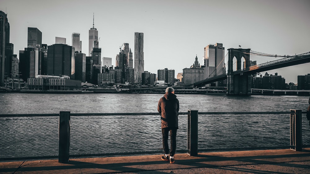 person standing and facing back while holding on railings viewing Brooklyn Bridge and Brooklyn city