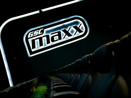 A row of empty black theater seats is shown beside a large illuminated sign displaying 'GSC maxx' against a dark background.