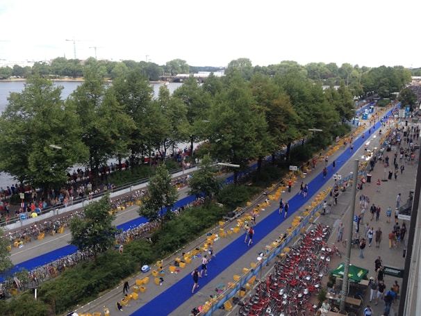 An aerial view of a busy outdoor event featuring a long blue carpeted path lined with bicycles and crowds of people on both sides. The scene is situated beside a body of water, with a row of green trees separating the path from the waterfront. People appear to be walking or running along the paths, and more bicycles are parked in designated areas. The area is bustling with activity, with many people gathered around.