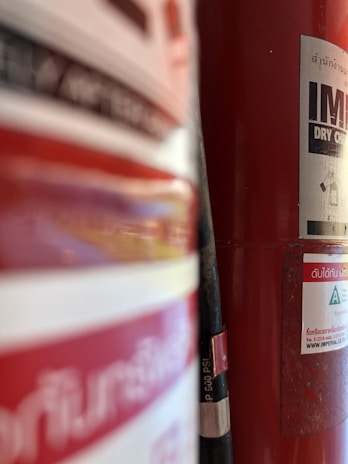 A close-up view of a red fire extinguisher with partially visible labels, hose, and some text in a foreign language. The background is slightly out of focus, emphasizing the fire extinguisher's details.
