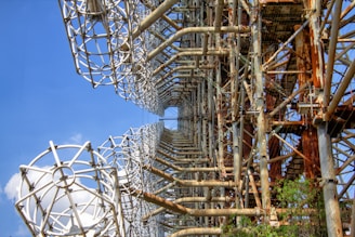 A large, intricate metal structure with an array of rusty beams and grid-like frameworks. The structure stands against a clear blue sky, showcasing a pattern of geometric shapes and cylindrical elements. The metal appears aged, with areas of rust highlighting its industrial design. The structure is surrounded by some greenery, suggesting an outdoor setting.