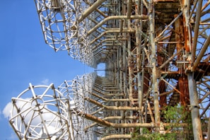 A large, intricate metal structure with an array of rusty beams and grid-like frameworks. The structure stands against a clear blue sky, showcasing a pattern of geometric shapes and cylindrical elements. The metal appears aged, with areas of rust highlighting its industrial design. The structure is surrounded by some greenery, suggesting an outdoor setting.