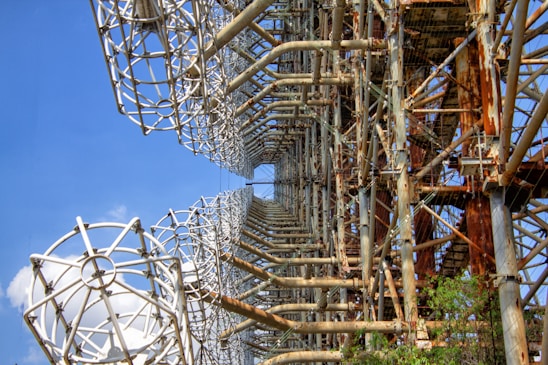 A large, intricate metal structure with an array of rusty beams and grid-like frameworks. The structure stands against a clear blue sky, showcasing a pattern of geometric shapes and cylindrical elements. The metal appears aged, with areas of rust highlighting its industrial design. The structure is surrounded by some greenery, suggesting an outdoor setting.
