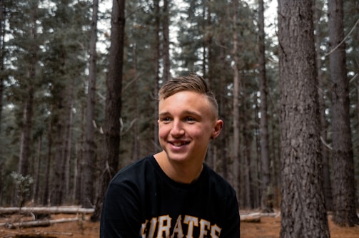 A young person with short hair is smiling while standing in a forest. They are wearing a dark t-shirt with the word 'PIRATES' visibly printed on it. The background consists primarily of tall trees and a forest floor covered in pine needles.
