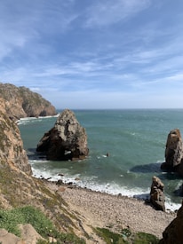 A rocky coastline with steep cliffs towering over a sandy and pebbled beach. The sea is a vibrant shade of blue-green with small waves crashing against the rocks. Above, the sky is mostly clear with the presence of some light clouds. Large rock formations jut out from the water, adding to the dramatic landscape.