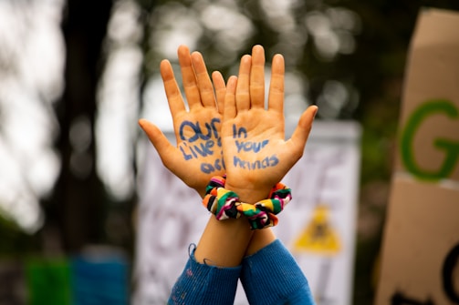 Two hands are raised in front of a blurred outdoor background, with the message 'Our lives in your hands' written in blue ink on the palms. Brightly colored scrunchies are worn on the wrists.