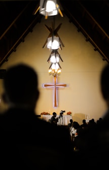 A church interior with a large wooden cross prominently mounted on the wall in the background. The setting is dimly lit, with several pendant lights hanging from the high ceiling casting soft light. People are seated in pews, facing the cross, and a man is standing with a camera, possibly recording an event.