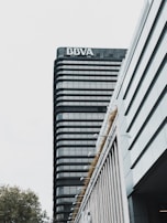A modern high-rise building with a prominent sign at the top. The building features a sleek design with dark glass windows and horizontal lines. The foreground includes another structure with vertical lines and greenery along its edges. The sky is overcast, creating a muted background.