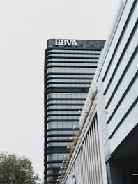 A modern high-rise building with a prominent sign at the top. The building features a sleek design with dark glass windows and horizontal lines. The foreground includes another structure with vertical lines and greenery along its edges. The sky is overcast, creating a muted background.