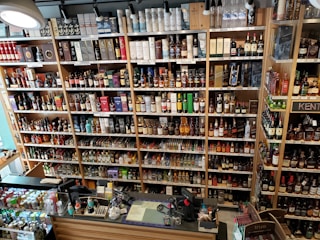 A well-organized liquor store shelf stacked with a variety of alcoholic beverages. Numerous bottles of whisky, wine, and spirits are displayed in rows, categorized by type and brand. The shelves are made of wood, providing a warm and inviting ambiance. The checkout counter is visible in the foreground, equipped with cash registers and various promotional materials.