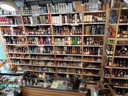 A well-organized liquor store shelf stacked with a variety of alcoholic beverages. Numerous bottles of whisky, wine, and spirits are displayed in rows, categorized by type and brand. The shelves are made of wood, providing a warm and inviting ambiance. The checkout counter is visible in the foreground, equipped with cash registers and various promotional materials.