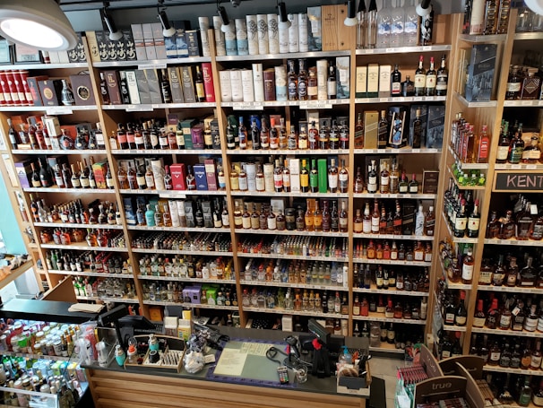 A well-organized liquor store shelf stacked with a variety of alcoholic beverages. Numerous bottles of whisky, wine, and spirits are displayed in rows, categorized by type and brand. The shelves are made of wood, providing a warm and inviting ambiance. The checkout counter is visible in the foreground, equipped with cash registers and various promotional materials.
