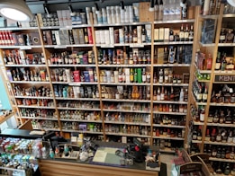 A well-organized liquor store shelf stacked with a variety of alcoholic beverages. Numerous bottles of whisky, wine, and spirits are displayed in rows, categorized by type and brand. The shelves are made of wood, providing a warm and inviting ambiance. The checkout counter is visible in the foreground, equipped with cash registers and various promotional materials.