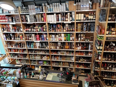 A well-organized liquor store shelf stacked with a variety of alcoholic beverages. Numerous bottles of whisky, wine, and spirits are displayed in rows, categorized by type and brand. The shelves are made of wood, providing a warm and inviting ambiance. The checkout counter is visible in the foreground, equipped with cash registers and various promotional materials.