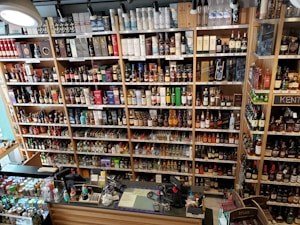 A well-organized liquor store shelf stacked with a variety of alcoholic beverages. Numerous bottles of whisky, wine, and spirits are displayed in rows, categorized by type and brand. The shelves are made of wood, providing a warm and inviting ambiance. The checkout counter is visible in the foreground, equipped with cash registers and various promotional materials.