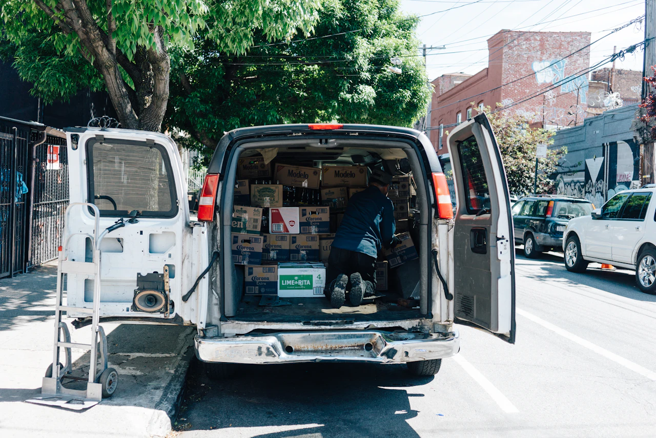 A driver loading cases into the back of a white delivery van at the curb, the vehicle inventory dispatch has to know before assigning an order