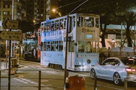 A bustling city street at night featuring a double-decker tram and a silver car. The tram has a blue advertisement on its side and several passengers visible inside. Bright streetlights illuminate the scene alongside a sign for Amoy Street. The background includes tall buildings and trees, giving a sense of urban life.