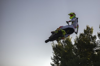 A motocross rider performs a high jump, mid-air against a backdrop of a clear sky and silhouetted trees. The rider wears a vibrant outfit with a neon helmet, and red and yellow accents. The scene captures dynamic movement and the thrill of extreme sports.