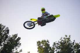 A motocross rider is performing a high jump on a dirt bike against a clear blue sky. The rider is wearing a helmet and protective gear, and the image captures the bike tilted in mid-air, showcasing a dynamic stunt. Trees are visible in the background.