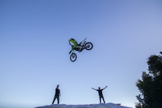 A motorcycle is airborne, performing a stunt high above a large inflated landing pad. Two people stand on the ground with raised arms, seemingly cheering or watching the stunt. The scene takes place under a clear blue sky with some trees visible to the side.