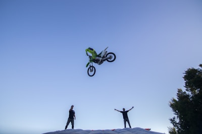 A motorcycle is airborne, performing a stunt high above a large inflated landing pad. Two people stand on the ground with raised arms, seemingly cheering or watching the stunt. The scene takes place under a clear blue sky with some trees visible to the side.
