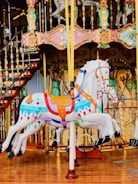 A colorful carousel with intricately designed horses featuring vibrant colors and ornate decorations. The image includes detailed carving work around the edges, displaying motifs of faces and floral patterns. The background shows elements of the carousel structure, including railings and decorative panels.
