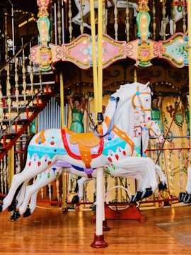 A colorful carousel with intricately designed horses featuring vibrant colors and ornate decorations. The image includes detailed carving work around the edges, displaying motifs of faces and floral patterns. The background shows elements of the carousel structure, including railings and decorative panels.