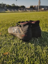 A pair of black football cleats with red stitching is placed on a lush green grass field. The shoes are positioned with the heels facing the viewer, revealing branding details. In the background, there is a blurry view of an empty sports stadium with rows of seating and large floodlights.