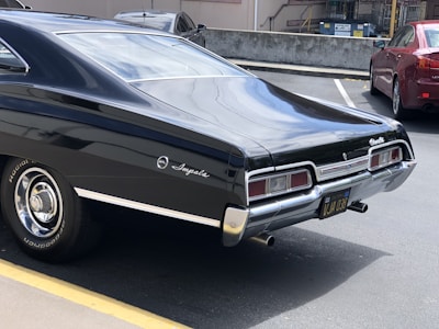 A classic black Chevrolet Impala is parked in a lot. The vehicle has a sleek, shiny exterior and features radial tires. The parking area is bordered by a yellow line, and other cars are visible in the background.