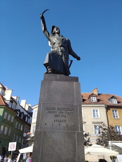 A tall bronze statue of a historical figure holding a sabre stands prominently on a pedestal. The inscription on the pedestal is in Polish, and around the statue are traditional European-style buildings with red-tiled roofs. The sky is clear and blue, casting a bright, natural light on the scene.
