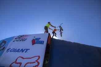 A person wearing a bright shirt is assisting another person who is launching off a ramp with a BMX bicycle. The scene is set against a clear blue sky, giving a sense of height and movement. Brand logos are visible on the ramp surface.