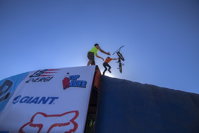 A person wearing a bright shirt is assisting another person who is launching off a ramp with a BMX bicycle. The scene is set against a clear blue sky, giving a sense of height and movement. Brand logos are visible on the ramp surface.