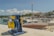 A coastal scene with a mechanical boat winch in the foreground, painted in blue and yellow, positioned on a concrete dock. A boat bearing the identifier OM 1359 is docked nearby. The background features a marina with several white boats moored in a calm harbor, and a row of buildings with red-tiled roofs along the waterfront. Trees, streetlights, and a few people walking can also be seen. The sky is clear with a few scattered clouds.