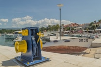 A coastal scene with a mechanical boat winch in the foreground, painted in blue and yellow, positioned on a concrete dock. A boat bearing the identifier OM 1359 is docked nearby. The background features a marina with several white boats moored in a calm harbor, and a row of buildings with red-tiled roofs along the waterfront. Trees, streetlights, and a few people walking can also be seen. The sky is clear with a few scattered clouds.