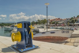 A coastal scene with a mechanical boat winch in the foreground, painted in blue and yellow, positioned on a concrete dock. A boat bearing the identifier OM 1359 is docked nearby. The background features a marina with several white boats moored in a calm harbor, and a row of buildings with red-tiled roofs along the waterfront. Trees, streetlights, and a few people walking can also be seen. The sky is clear with a few scattered clouds.