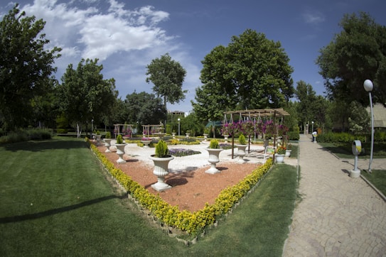 A well-maintained garden with a geometric arrangement of green grass, pathways, and flower arrangements. The central area features large white planters with greenery and a pergola adorned with hanging flowers. Tall, lush trees surround the garden, and a stone walkway intersects the scene on the right.