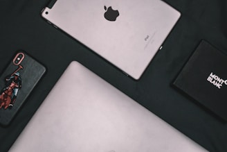 A flat lay composition of electronic devices and a box. A silver tablet with the Apple logo, an iPhone with a custom case featuring a polo player, a laptop with a metallic finish, and a black Montblanc box are arranged on a dark surface.