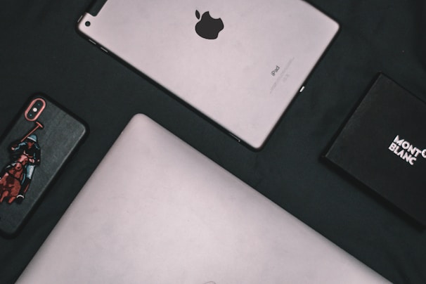 A flat lay composition of electronic devices and a box. A silver tablet with the Apple logo, an iPhone with a custom case featuring a polo player, a laptop with a metallic finish, and a black Montblanc box are arranged on a dark surface.