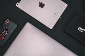 A flat lay composition of electronic devices and a box. A silver tablet with the Apple logo, an iPhone with a custom case featuring a polo player, a laptop with a metallic finish, and a black Montblanc box are arranged on a dark surface.