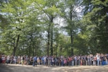A large group of people is gathered in a forested area, surrounded by tall green trees. The crowd includes individuals of varying ages, standing closely together. The atmosphere appears lively, with some participants taking photos or interacting with each other.