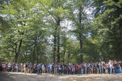 A large group of people is gathered in a forested area, surrounded by tall green trees. The crowd includes individuals of varying ages, standing closely together. The atmosphere appears lively, with some participants taking photos or interacting with each other.