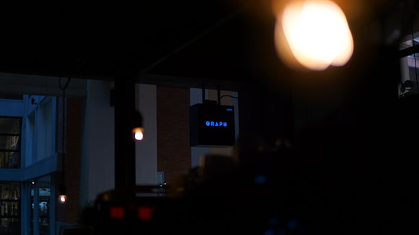 A dimly lit indoor environment with an illuminated 'GRAPH' sign in the background. Warm overhead lights create a cozy atmosphere, with a focus on the contrast between darkness and the bright sign.