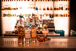 Three bottles of Una Vida tequila are displayed on a marble countertop, complemented by a bowl of fresh fruits and a cocktail. The background is filled with a well-lit bar shelf showcasing various bottles.