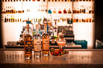 Three bottles of Una Vida tequila are displayed on a marble countertop, complemented by a bowl of fresh fruits and a cocktail. The background is filled with a well-lit bar shelf showcasing various bottles.