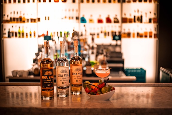 Three bottles of Una Vida tequila are displayed on a marble countertop, complemented by a bowl of fresh fruits and a cocktail. The background is filled with a well-lit bar shelf showcasing various bottles.